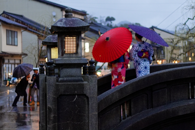 Women in yukata with traditional umbrellas crossing a stone bridge on a rainy day in Kinosaki Onsen, capturing its timeless charm.