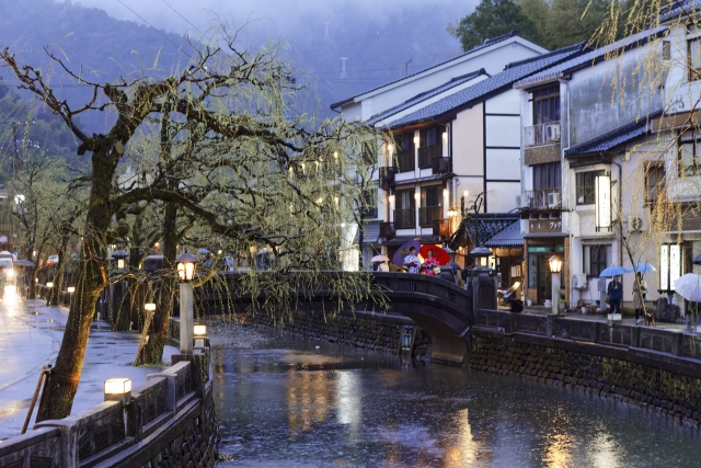 Rainy evening in Kinosaki Onsen with willow-lined canal, traditional ryokan, and visitors crossing the bridge with colorful umbrellas.