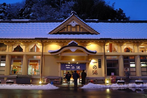 Front view of Ichinoyu public bath covered in snow at night in Kinosaki Onsen, glowing warmly against the winter backdrop.