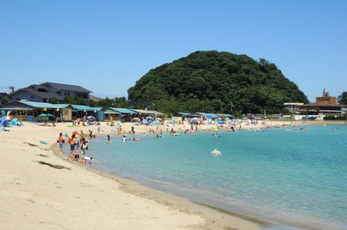 Visitors enjoying the sunny beach near Dogo Onsen, with clear blue waters and lush green hills in the background.