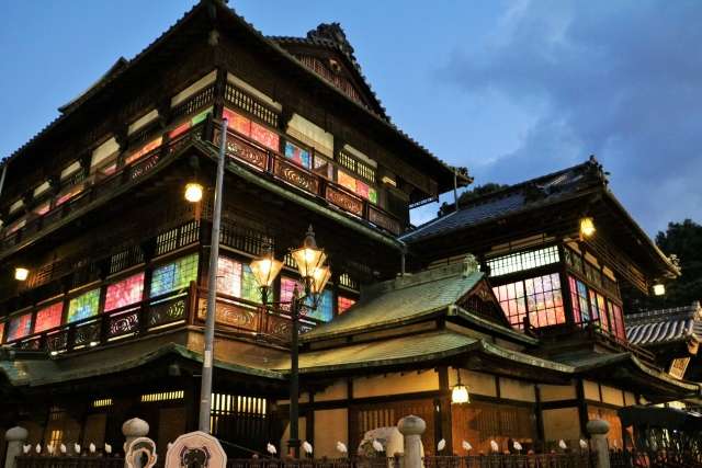 A night view of Dogo Onsen Honkan in Matsuyama, with its stained-glass-like windows illuminated in colorful lights, highlighting the traditional wooden architecture.