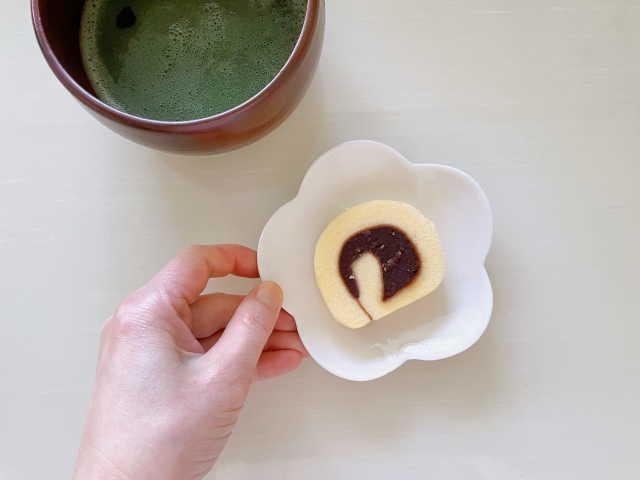 A hand serving a slice of Dogo Roll with light sponge and red bean paste, paired with a cup of matcha — a refined tea-time moment in Dogo Onsen.