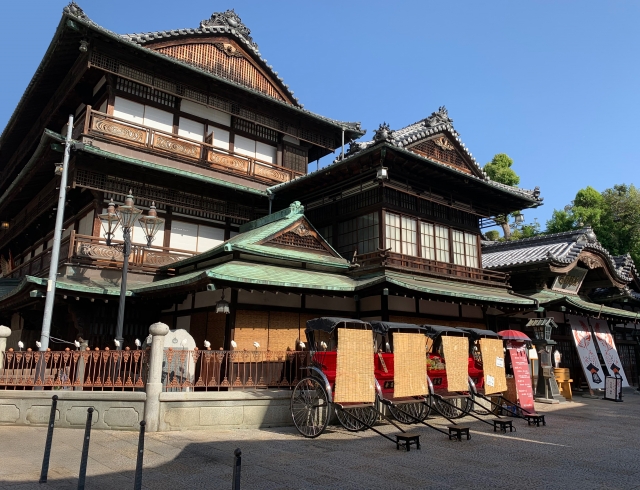 The historic Dogo Onsen Honkan building in Matsuyama, with traditional rickshaws lined up in front under a clear blue sky.