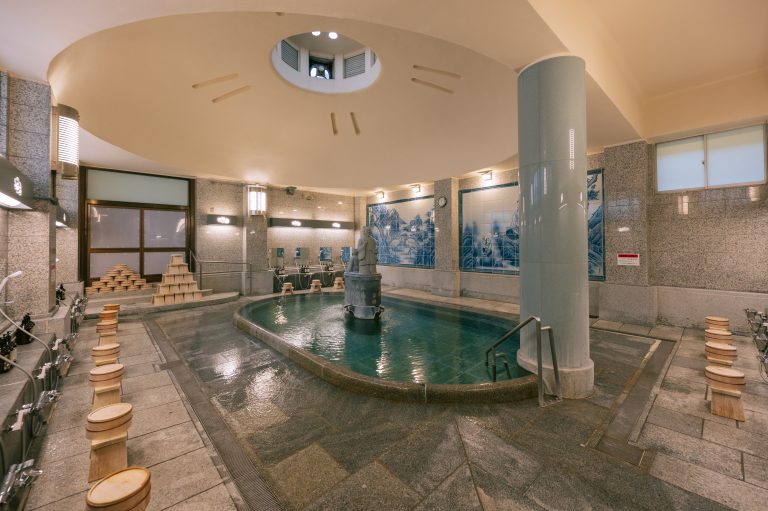 Spacious Kami-no-Yu bath in Dogo Onsen Honkan, featuring a central fountain, tiled murals, and traditional washing stations.