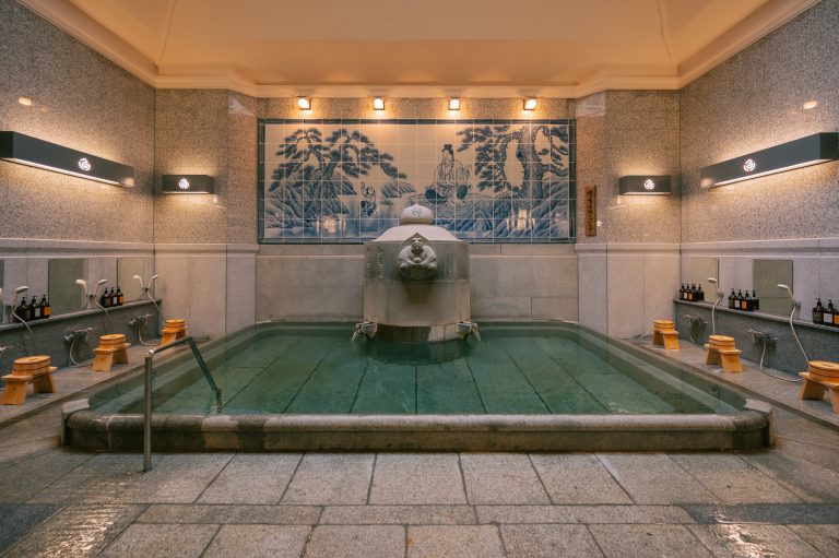 Elegant indoor bath in Dogo Onsen featuring a central fountain and a blue tile mural depicting the legend of Prince Shōtoku and a white heron.