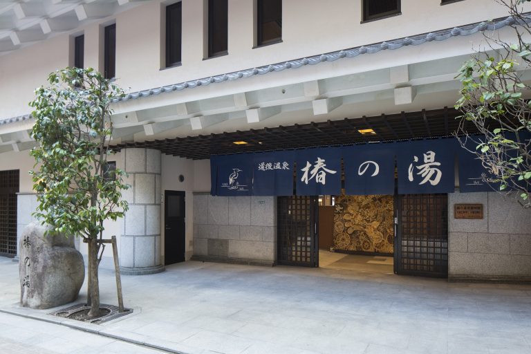 Entrance to Tsubaki-no-Yu, a modern facility of Dogo Onsen, featuring traditional Japanese design and a bold blue curtain with white calligraphy.