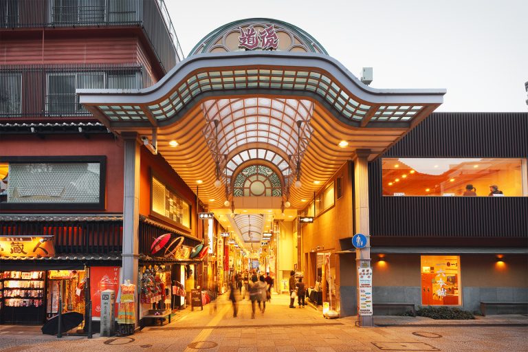 A lively shopping arcade entrance in Dogo Onsen with traditional storefronts and illuminated signs, capturing the local atmosphere at dusk.