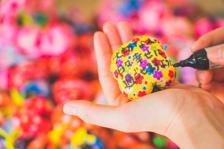 A person writing a wish on a colorful, flower-patterned omamori charm in Dogo Onsen, Japan.