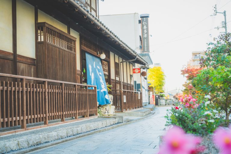 A charming traditional wooden building with a blue noren curtain along a quiet street in Dogo Onsen, framed by colorful seasonal flowers.