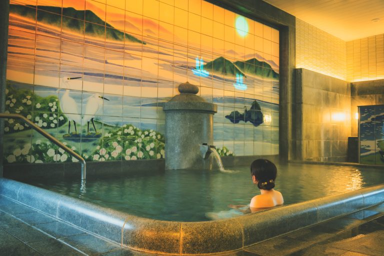 A woman relaxing in the indoor bath of Dogo Onsen Asuka-no-Yu, with a mural depicting herons and a serene coastal landscape.