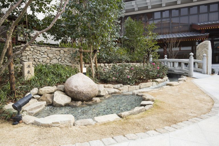 The original hot spring source of Dogo Onsen, featuring a large round stone surrounded by a small stream, garden plants, and a stone bridge.