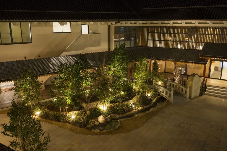 Night view of the illuminated courtyard garden at Dogo Onsen Annex Asuka-no-Yu, featuring lush greenery and a small arched bridge.