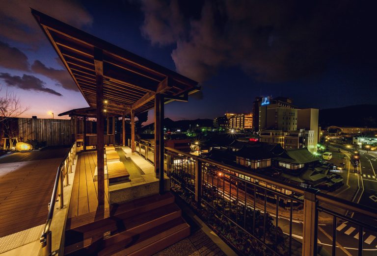 Night view from the rooftop foot bath of Dogo Onsen, overlooking the illuminated townscape and traditional bathhouse buildings under a dramatic evening sky.