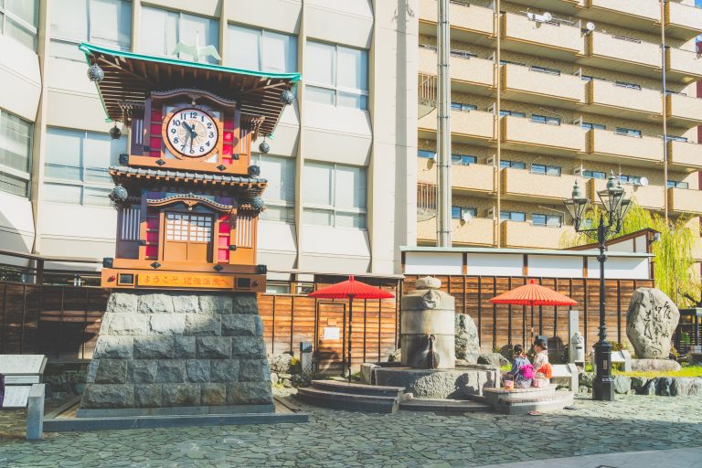 Botchan Karakuri Clock and footbath area in front of Dogo Onsen Station, where visitors relax under red parasols in a welcoming public space.