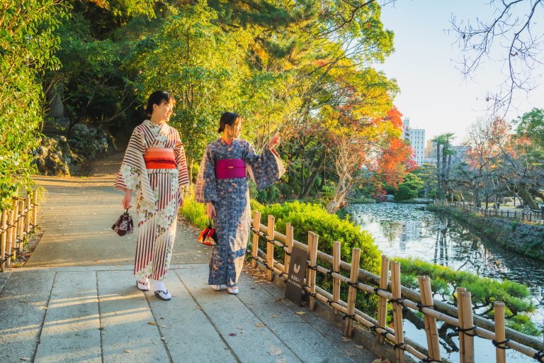 Two women in colorful yukata strolling along a scenic riverside path surrounded by autumn foliage in Dogo Onsen, Japan.