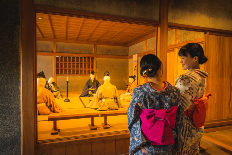 Two women in yukata observing a historical reenactment with mannequins dressed as samurai inside a traditional Japanese room in Dogo Onsen.